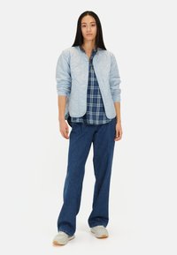 Lyseblå quiltet jakke over en ternet skjorte, parret med brede denimbukser og lyse sneakers. Sættet har bløde teksturer og et afslappet design.