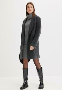 Manteau en laine gris foncé avec des revers crantés, des boutons doubles et des poches latérales. Porté par-dessus une robe pull gris et des bottes noires à hauteur de genou.