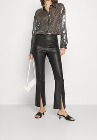 Camicia nera con paillettes e bottoni, pantaloni in pelle svasati con spacchi, e mule nere con tacco. Una borsa bianca completa l'ensemble.
