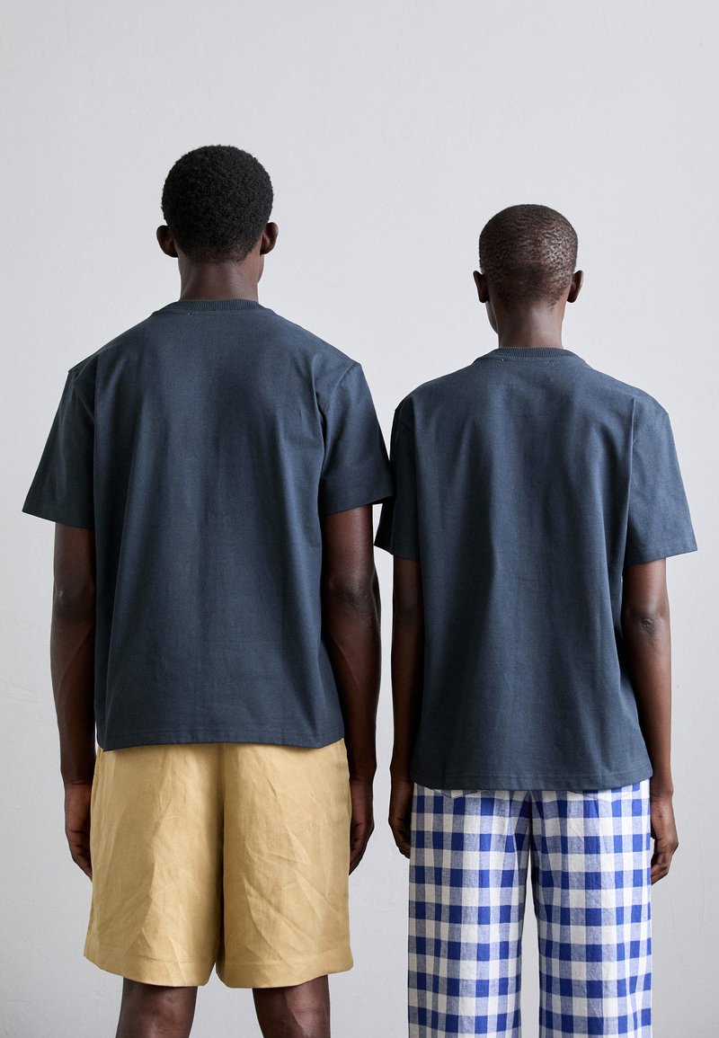 Two individuals stand back-to-back, wearing dark short-sleeved T-shirts. One wears beige shorts, the other blue and white checkered trousers.