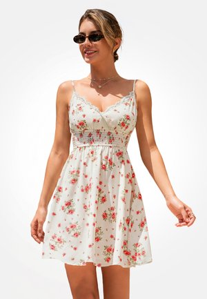 Jonge vrouw met zonnebril en een witte mouwloze jurk met rode bloemenprint, glimlachend tegen een effen achtergrond.