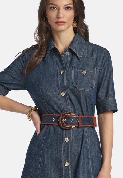 Femeie purtând o rochie din denim cu nasturi, mâneci scurte, buzunar frontal la piept și o curea lată cu o cataramă mare.