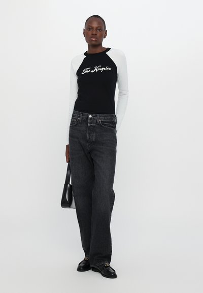 Camisa de manga larga en blanco y negro con texto, combinada con unos pantalones vaqueros negros de cintura alta. Complementada con un pequeño bolso negro y zapatos negros.