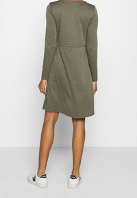 Długa, oliwkowa sukienka z długim rękawem, dopasowanym gorsetem i luźną spódnicą, z okrągłym dekoltem i bocznymi rozcięciami. Noszona z białymi sneakersami.