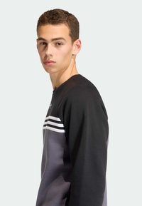 Čierna a sivá mikina s okrúhlym výstrihom, dlhými rukávmi a horizontálnymi bielymi pruhmi na hrudi. Mäkká textúra látky.