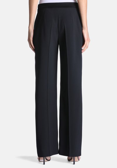 Femme portant un pantalon noir taille haute à jambes larges et des sandales noires à talons hauts, vue de dos sur un fond uni.
