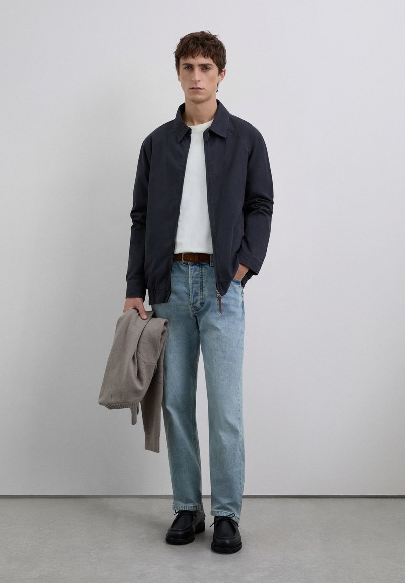 Jeune homme portant une veste bleu marine, un t-shirt blanc, un jean bleu clair, des chaussures noires, tenant un pull beige contre un fond clair uni.