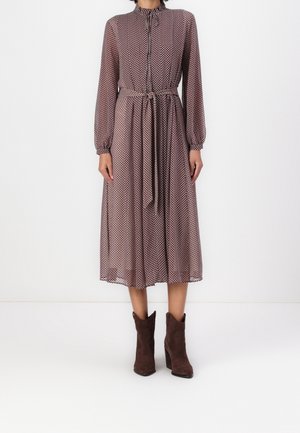 Woman wearing a long-sleeve, brown midi dress with small white polka dots, a tied waist, and brown heeled ankle boots.
