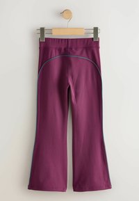 Pantalon ajusté et évasé couleur bordeaux avec une texture lisse, présentant des surpiqûres contrastantes bleues le long des coutures latérales. Suspendu sur un cintre en bois.