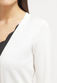 White knitted cardigan with a V-neck design. Features a soft texture and fitted cut, partially covering a black top underneath.