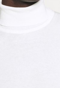 Primo piano di una persona che indossa un maglione bianco a collo alto, che mostra il collo e la parte superiore del petto.
