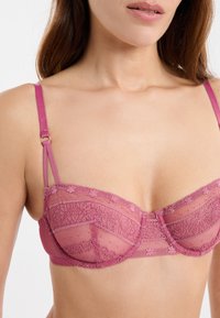 Różowy koronkowy biustonosz z przejrzystymi miseczkami z siateczki, kwiatowymi wzorami, regulowanymi ramiączkami i delikatnym wykończeniem. Złote elementy ozdobne.