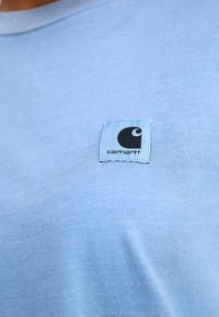 Světle modré bavlněné tričko s kulatým výstřihem a černou visačkou Carhartt na levé části hrudi. Měkká textura a minimalistický design.