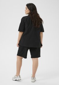 Chemise noire à manches courtes et shorts noirs en tissu léger. Le modèle porte des baskets blanches. Design simple et décontracté.