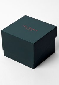 Ciemnozielone, sztywne pudełko kartonowe z klapą na górze; na wieczku, na środku, znajduje się napis "TED BAKER LONDON" w kolorze czerwonym; gładka tekstura.