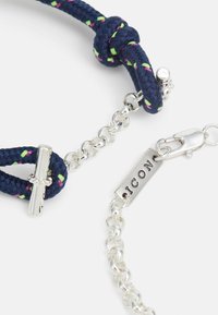 Bracelet en corde tressée marine avec des accents roses et verts, présentant une chaîne en argent, un fermoir et une étiquette de marque.