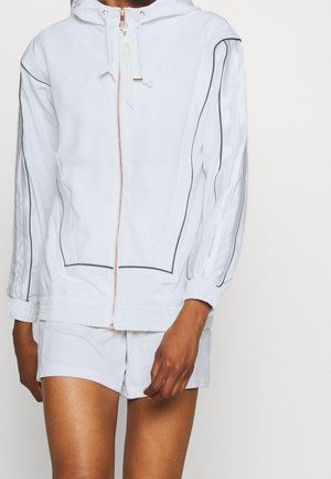 Persona con chaqueta blanca con capucha y cremallera, con ribetes negros y pantalones cortos blancos a juego, de pie frente a un fondo claro y liso.