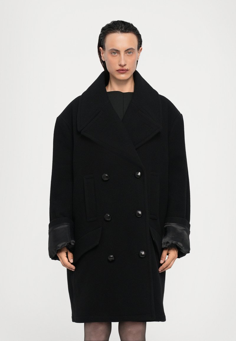 Black wool peacoat with large lapels, double-breasted button closure, side pockets, and satin-lined cuffs. Minimalist design, mid-length.