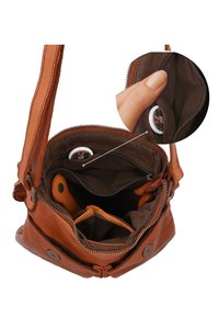 Bruine leren crossbodytas open om binnenvakken en een klein wit rond voorwerp in een ritssluitingvak te tonen, vastgehouden door een hand.