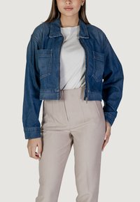 Giubbotto di jeans ritagliato di blu medio con due tasche frontali, chiusura con zip e spalle strutturate, abbinato a pantaloni beige chiaro.