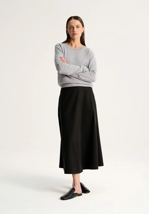 Kobieta stoi ze skrzyżowanymi ramionami, ubrana w szary sweter, czarną spódnicę midi i czarne buty wsuwane na tle jednolitego jasnego tła.