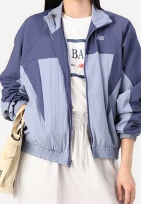 Žena v modré a levandulové zipové bundě nad bílým tričkem s potiskem, držící béžovou kabelku, s letní sukní světlé barvy.