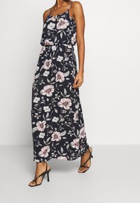 Robe maxi florale avec un fond bleu marine, ornée de fleurs roses et blanches, avec des fines bretelles et une coupe ample. Assortie à des sandales noires à talons hauts.