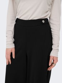 Pantalones negros con una textura suave, con un detalle de botón en la cintura, combinados con una camiseta de manga larga de color beige claro.