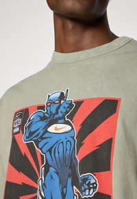 Grøn bomulds T-shirt med et blåt grafik af en muskuløs karakter på en rød og sort burst baggrund, med et Nike-logo i øverste venstre hjørne.
