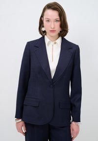 Blazer tailored color navy con rever a taglio, due tasche frontali e dettagli di cucitura blu discreti. Abbinato a una blusa crema con apertura a chiave.