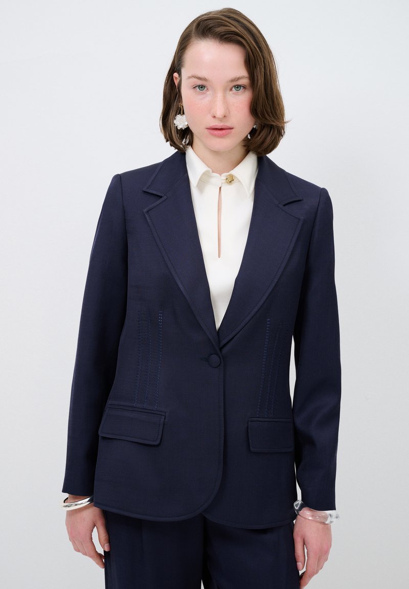 Blazer tailored color navy con rever a taglio, due tasche frontali e dettagli di cucitura blu discreti. Abbinato a una blusa crema con apertura a chiave.