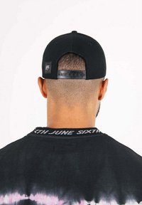 Cappellino nero con chiusura snapback e una piccola etichetta con logo, abbinato a una maglietta nera tie-dye con scritte bianche sul colletto.