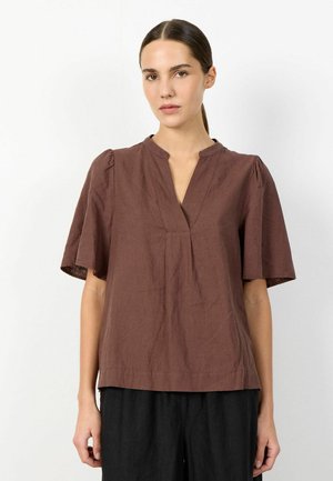 Nő barna V-nyakú, rövid ujjú blúzban és fekete nadrágban, sima fehér háttér előtt állva.