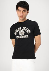 Camiseta de algodón negra con cuello redondo, que presenta un prominente estampado gráfico en blanco y dorado "Guess Jeans California" en el frontal.