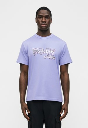 Tânăr care poartă un tricou lavandă Versace Jeans Couture și pantaloni negri, stând pe un fundal alb simplu.