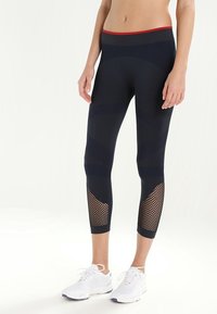 Marinblå leggings i texturerad tyg, med nätpaneler vid underbenen och en kontrasterande röd midjebanddetalj.