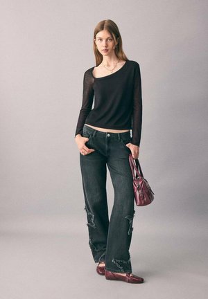 Donna in un top lungo asimmetrico nero a maniche lunghe, jeans neri strappati, ballerine bordeaux, e che tiene una borsa bordeaux su uno sfondo grigio.