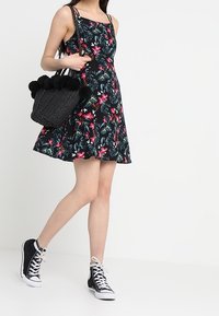 Sukienka bez rękawów w kwiatowy wzór z dopasowanym topem i rozkloszowaną spódnicą, na czarnym tle; zestawiona z teksturowaną czarną torbą i sneakersami za kostkę.