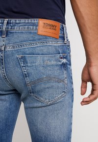 Blå denimjeans med en ljus tvätt, med en läderetikett som säger "TOMMY JEANS", och kontrasterande sömmar på bakfickorna.