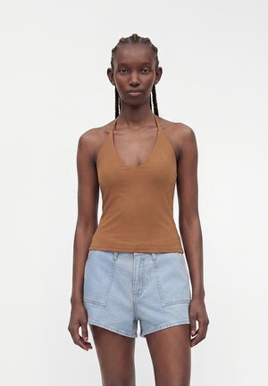 Femme aux cheveux tressés portant un haut marron à col halter et un short en denim bleu clair, debout devant un fond blanc uni.