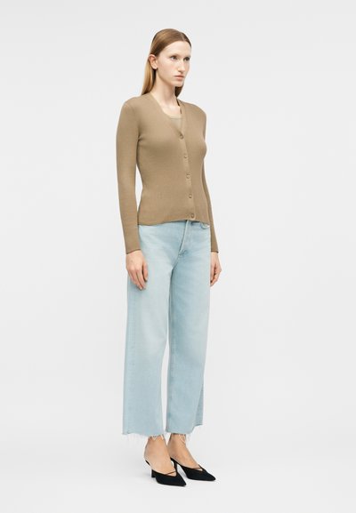 Γυναίκα όρθια, φοράει ένα μπεζ ζακέτα με κουμπιά, ανοιχτό μπλε τζιν με φαρδιά πόδια και φθαρμένα τελειώματα, και μαύρες μυτερές γόβες.