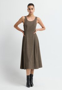 Mujer de pie con las manos en las caderas, vistiendo un vestido midi sin mangas a cuadros marrones y botas negras hasta el tobillo, frente a un fondo blanco liso.