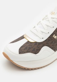 Witte en bruine sneaker met een gestructureerd bovenwerk van bruin leer, gouden accenten, witte veters en een rubberen zool met een geribbeld ontwerp.