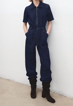 Personne portant une combinaison en jean bleu foncé avec fermeture éclair devant, ceinture, manches courtes et bottes noires souples, debout contre un mur blanc.