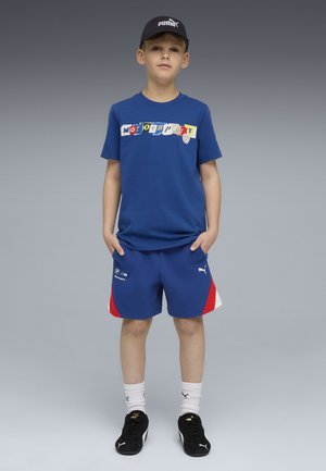 Dreng står med hænderne i lommerne iført blå motorsport t-shirt, matchende shorts med røde accenter, sort Puma kasket og sorte sneakers.