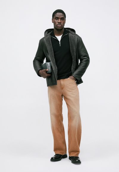 Schwarze Lederjacke mit fellgefütterter Kapuze, schwarzer Pullover, hellbraune gerade Jeans und schwarze Schuhe. Hält eine kleine schwarze Tasche.