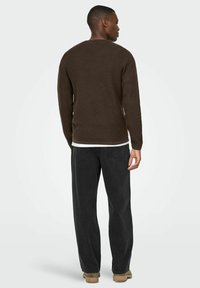 Pull en maille marron foncé à manches longues, col rond et texture côtelée, associé à un jean noir à coupe droite et des chaussures beiges.