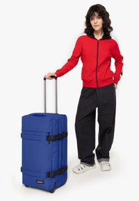 Modrý cestovní kufr s texturovaným povrchem, nastavitelnou teleskopickou rukojetí, černými popruhy a koly. Doprovázen osobou v červené bundě.