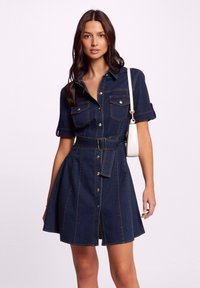 Tummansinisestä denim-mekosta, jossa on lyhyet hihat, nappilista edessä, kaksi rintataskua, vyötäröltä kapeaksi muotoutuva malli ja liehuva helma.