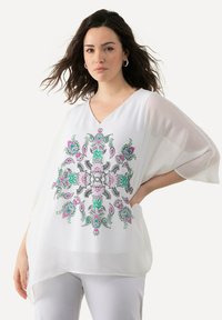 Blusa bianca larga con scollatura a V, caratterizzata da ricami floreali colorati in verde e rosa, tessuto trasparente e maniche ampie.